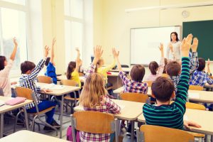 education, elementary school, learning and people concept - group of school kids with teacher sitting in classroom and raising hands